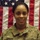 A U.S. Army soldier named Gordon is photographed in an official portrait-style image in front of an American flag. She is wearing the Army Combat Uniform (ACU) in MultiCam/OCP pattern with name tape 'GORDON' and 'U.S. ARMY' branch tape visible. The subject is smiling and appears to be posing for an official or semi-official military photograph.