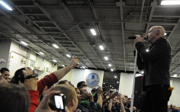 A bald male performer is singing into a microphone on a raised stage inside what appears to be a military vessel's hangar bay, indicated by the official seal visible on the wall. A large crowd of audience members, many holding up phones and cameras to record the performance, are gathered in front of the stage. The setting suggests this is a USO-style entertainment event for military personnel.