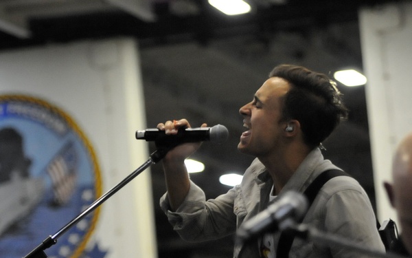 A male performer is singing into a handheld microphone on stage, wearing a gray jacket and an in-ear monitor. The performance appears to be taking place at a military venue, as evidenced by a large official military unit insignia/seal visible on the wall behind him. A second person, appearing bald, is partially visible in the foreground right.