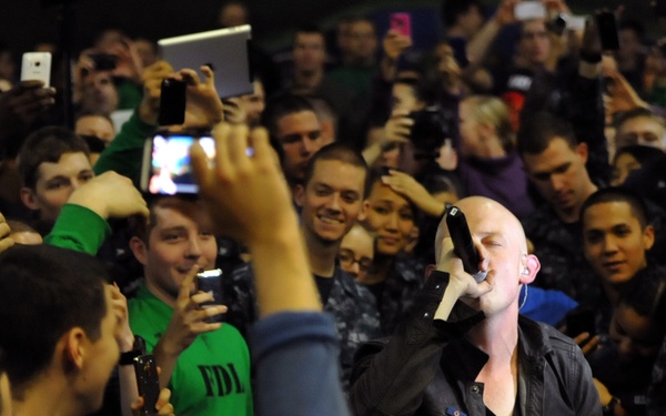 A bald male performer is singing into a microphone while surrounded by a large, enthusiastic crowd at what appears to be an indoor concert or live event. The audience is densely packed around the performer, many holding up smartphones and tablets to capture the moment. The intimate setting suggests the performer is in the middle of the crowd rather than on a traditional stage.