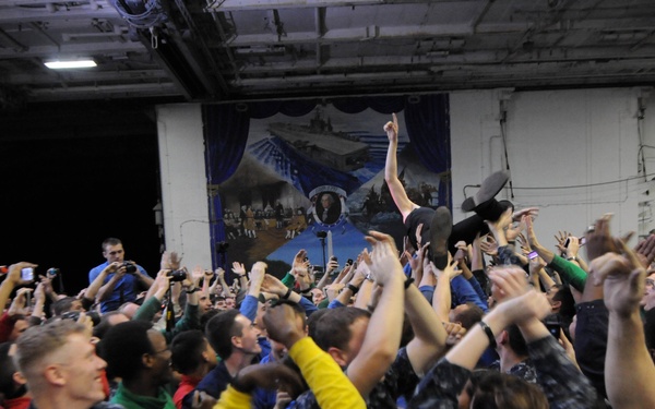 A large crowd of people, many wearing U.S. Navy camouflage uniforms, are gathered in what appears to be a ship's hangar bay during a celebratory event. The crowd is energetic with arms raised, phones and cameras held up, and expressions of excitement. A large decorative banner or crest is visible on the wall in the background.