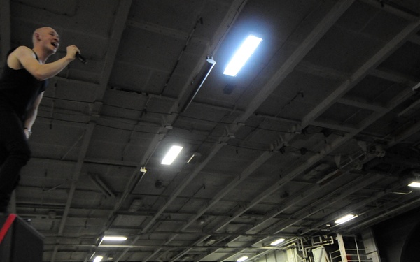 A bald performer stands elevated on top of a speaker stack, singing into a microphone while leaning toward a large crowd below. The concert appears to be taking place inside a military vessel, likely an aircraft carrier hangar bay, as indicated by the industrial ceiling structure and a visible banner reading 'CVT-70'. The crowd is densely packed and many audience members are holding up cameras and phones to capture the performance.