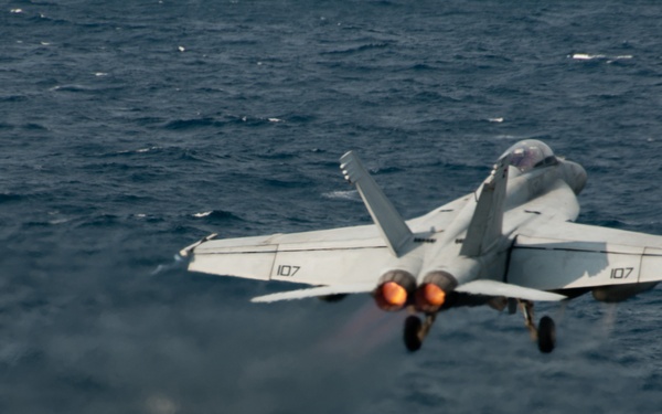 A U.S. Navy fighter jet, marked with the number '107' on both wings, is captured mid-launch from an aircraft carrier deck over open ocean. The aircraft's twin engines are visibly in afterburner, producing bright orange flame. Deck crew members in green jerseys are visible on the right side of the carrier deck.