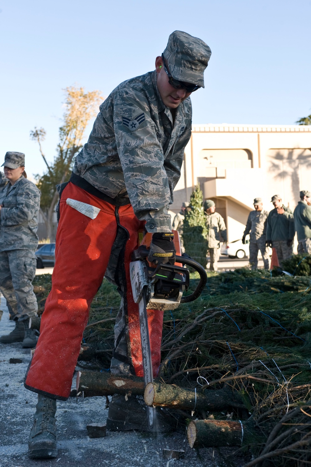 A U.S. Air Force service member in ABU (Airman Battle Uniform) is operating a chainsaw to cut through a log or tree trunk, wearing orange chainsaw safety chaps and protective eyewear. Several other uniformed military personnel are visible in the background observing the activity. The setting appears to be an outdoor area near a building, with cut branches and debris scattered on the ground.