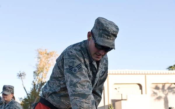 A U.S. Air Force service member in ABU (Airman Battle Uniform) is operating a chainsaw to cut through a log or tree trunk, wearing orange chainsaw safety chaps and protective eyewear. Several other uniformed military personnel are visible in the background observing the activity. The setting appears to be an outdoor area near a building, with cut branches and debris scattered on the ground.