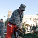 A U.S. Air Force service member in ABU (Airman Battle Uniform) is operating a chainsaw to cut through a log or tree trunk, wearing orange chainsaw safety chaps and protective eyewear. Several other uniformed military personnel are visible in the background observing the activity. The setting appears to be an outdoor area near a building, with cut branches and debris scattered on the ground.