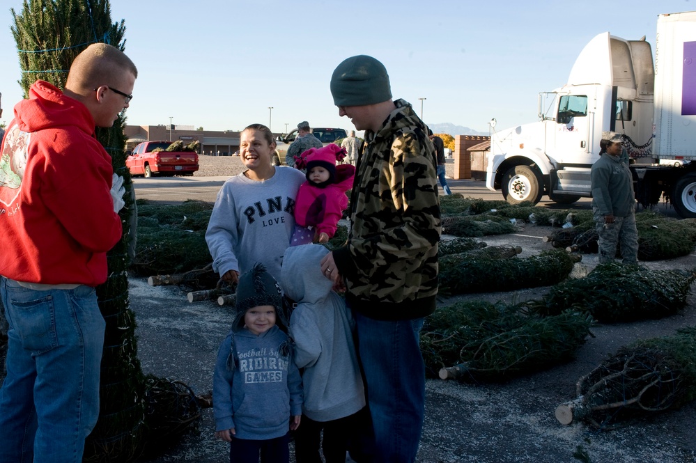 A family with young children is selecting a Christmas tree at an outdoor tree lot, likely on or near a military base given the uniformed personnel visible in the background. The group includes two adults, a toddler, an older child, and an infant dressed in pink. The atmosphere appears cheerful and festive, with cut Christmas trees laid out across the ground.