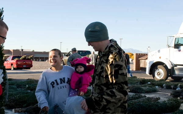 A family with young children is selecting a Christmas tree at an outdoor tree lot, likely on or near a military base given the uniformed personnel visible in the background. The group includes two adults, a toddler, an older child, and an infant dressed in pink. The atmosphere appears cheerful and festive, with cut Christmas trees laid out across the ground.