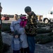 A family with young children is selecting a Christmas tree at an outdoor tree lot, likely on or near a military base given the uniformed personnel visible in the background. The group includes two adults, a toddler, an older child, and an infant dressed in pink. The atmosphere appears cheerful and festive, with cut Christmas trees laid out across the ground.