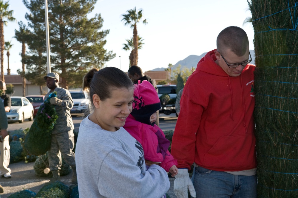 A family consisting of a woman holding a young child in pink clothing and a man in a red hoodie are examining Christmas trees at an outdoor tree lot. In the background, a person in military camouflage uniform is visible carrying a wreath or tree. The setting appears to be a parking lot in a warm-climate region, evidenced by palm trees and mountains in the background.