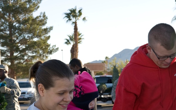 A family consisting of a woman holding a young child in pink clothing and a man in a red hoodie are examining Christmas trees at an outdoor tree lot. In the background, a person in military camouflage uniform is visible carrying a wreath or tree. The setting appears to be a parking lot in a warm-climate region, evidenced by palm trees and mountains in the background.