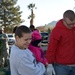 A family consisting of a woman holding a young child in pink clothing and a man in a red hoodie are examining Christmas trees at an outdoor tree lot. In the background, a person in military camouflage uniform is visible carrying a wreath or tree. The setting appears to be a parking lot in a warm-climate region, evidenced by palm trees and mountains in the background.