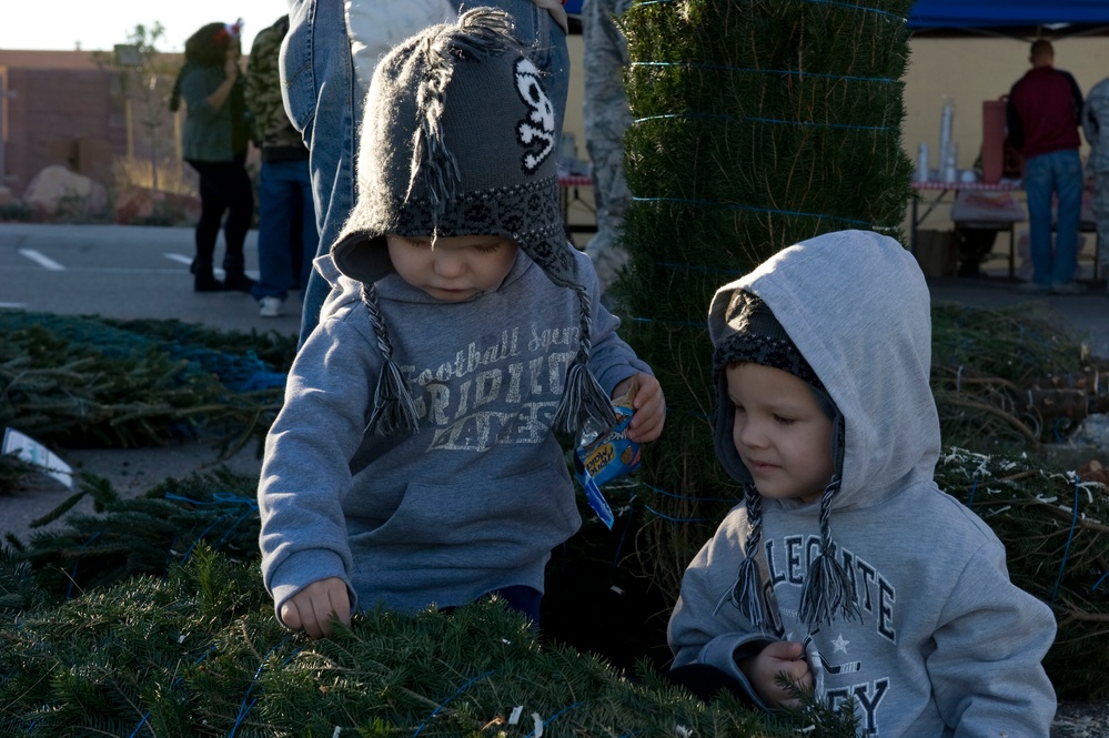 Two young toddlers wearing matching gray knit hats with skull designs and gray hooded sweatshirts are exploring cut Christmas trees at what appears to be an outdoor Christmas tree lot. The children are touching and examining the pine branches on the ground. Adults and a vendor tent are visible in the background.