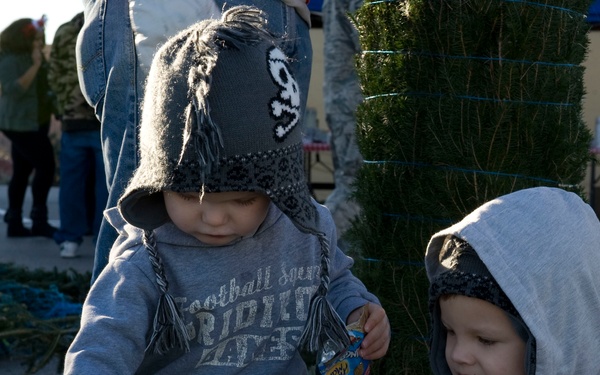 Two young toddlers wearing matching gray knit hats with skull designs and gray hooded sweatshirts are exploring cut Christmas trees at what appears to be an outdoor Christmas tree lot. The children are touching and examining the pine branches on the ground. Adults and a vendor tent are visible in the background.