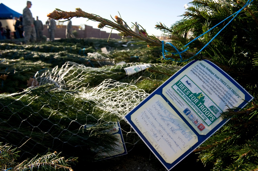 The image shows bundled Christmas trees tagged with 'Trees for Troops' program cards, a charitable initiative that ships real Christmas trees to deployed military personnel. A handwritten note is visible on the tag expressing holiday wishes and gratitude to service members. Military personnel in uniform are visible in the blurred background.