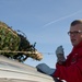 A young man wearing a red hoodie and glasses is securing a Christmas tree to the roof of a silver vehicle. He is wearing work gloves and appears to be tying or adjusting rope to fasten the bundled evergreen tree. The scene takes place outdoors under a clear blue sky.