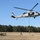A military helicopter, appearing to be an MH-60 Seahawk, hovers over an open field while personnel conduct fast-rope insertion/extraction (FRIES) training. One soldier is actively descending the rope while others are grouped on the ground in a security posture. The setting appears to be a military training area surrounded by pine trees under clear blue skies.
