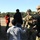 A group of civilians, including a child, are interacting with fully equipped military personnel in tactical gear at what appears to be an outdoor military demonstration or open day event. The soldiers are wearing camouflage uniforms, tactical vests, helmets, and carrying weapons, suggesting they are special operations or combat-ready units. The setting appears to be a military base or training facility with open fields and tree lines in the background.