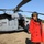 A smiling woman wearing a red coat and a navy captain's hat poses in front of a large gray military helicopter on an open grassy field. The helicopter displays a U.S. military star insignia on its fuselage and has its side door open. The setting appears to be a military airfield or open landing area with trees visible in the background.