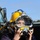 A woman wearing a purple jacket is holding a large professional diving helmet up to her face outdoors. The helmet is yellow and chrome/silver in color, appearing to be a Kirby Morgan brand commercial diving helmet. The setting appears to be a sunny outdoor location with a vehicle visible in the background.