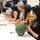 A group of individuals, likely celebrities or athletes, are seated at a long table participating in an autograph signing session. The woman in the foreground is signing a military-style olive drab combat helmet with a black marker. The setting appears to be a military base or event, suggested by the camouflage uniform visible on the left side of the image.