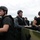 Several individuals wearing black tactical vests and helmets are seated aboard what appears to be a military-style watercraft on a body of water. They appear alert and are facing different directions, with at least one firearm visible resting on the vessel's edge. The setting and equipment suggest a tactical training exercise, law enforcement operation, or a film/television production.