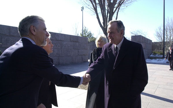 Two men in dark formal overcoats are shaking hands outdoors at what appears to be the Franklin D. Roosevelt Memorial in Washington, D.C., as indicated by the engraved text visible on the granite wall in the background. A woman in a black coat stands nearby observing the greeting, while another woman with blonde hair stands further back. The scene appears to be a formal or ceremonial visit during winter, evidenced by bare trees and patches of snow on the ground.