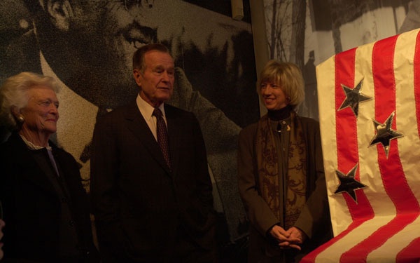 Three individuals are standing together in what appears to be a museum or memorial setting, posing near a large American flag with metallic star decorations. A large black-and-white photographic mural serves as the backdrop. The setting suggests a formal commemorative or dedication event.
