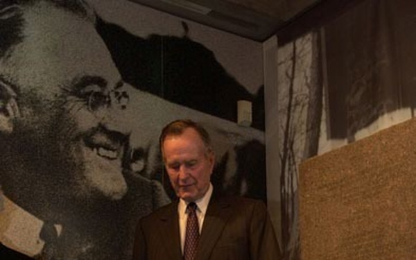 An elderly man in a dark suit and tie stands in what appears to be a museum or exhibition space, looking down at a glass display case or lectern. Behind him is a large black-and-white photographic mural of a smiling man wearing glasses. The setting suggests a formal visit to a commemorative or historical exhibit.