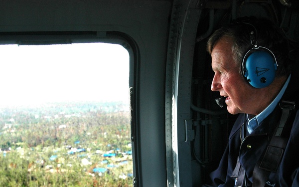 An older man wearing blue aviation headphones and a dark jacket is seated inside a helicopter, looking out a large window at a landscape below. The aerial view shows what appears to be a storm-damaged area with scattered blue tarps visible among trees and structures. A second person is partially visible on the left side of the frame.