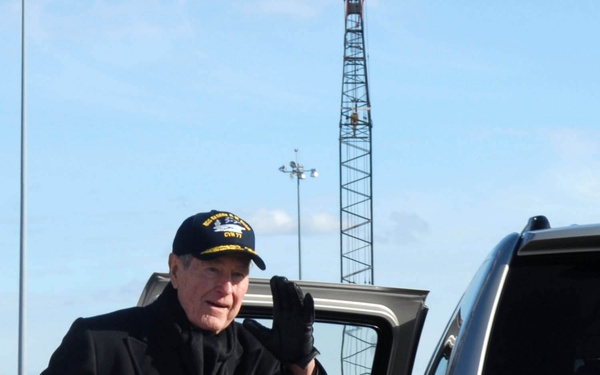 An elderly man dressed in a dark overcoat and black gloves is waving while entering or exiting a silver Chevrolet Suburban SUV. He is wearing a navy-style cap with visible text reading 'CVN 77'. The setting appears to be an outdoor area, possibly a naval facility or shipyard, with a crane and light towers visible in the background.