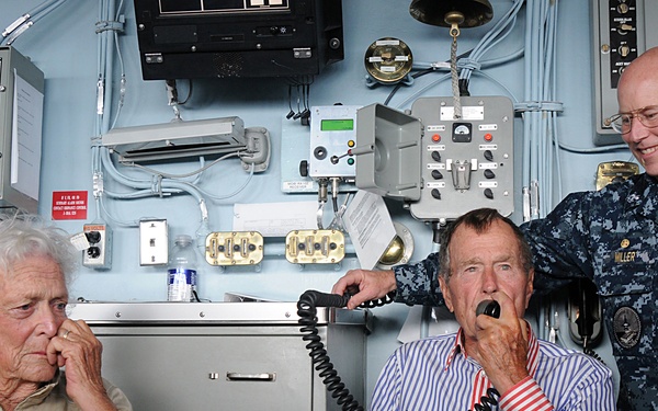 Three individuals are seated and standing inside what appears to be a naval vessel's control or communications room. A U.S. Navy officer in digital camouflage uniform stands to the right, while two elderly civilians are seated. The man in the center is speaking into a corded handset/telephone while the woman to his left listens.