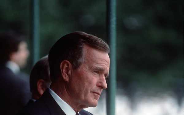 A middle-aged to older man in a dark navy suit and patterned red tie is seated in a burgundy/maroon chair, appearing to observe something off-camera. His hands are clasped in his lap and he has a composed, attentive expression. The background shows blurred greenery and at least one other person in formal attire standing behind him.
