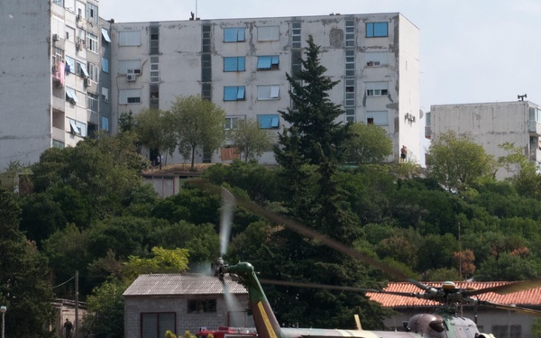 A Slovak military helicopter, identified as a Mi-17 SAR (Search and Rescue) variant, is on the ground with rotors spinning while several soldiers in combat gear run toward or away from it in an urban environment. The setting appears to be a post-Soviet style residential area with large concrete apartment blocks visible in the background. This appears to be a military exercise or training operation.