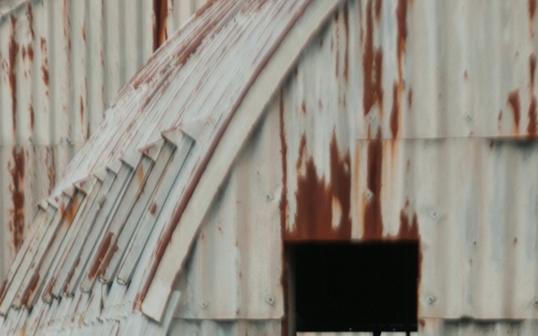 A silhouetted figure in full tactical/military gear stands in the doorway of a corrugated metal Nissen or Quonset hut showing significant rust and weathering. The figure appears to be equipped with a helmet, body armor, and what appears to be a weapon or equipment pack. The backlit composition renders the figure entirely in silhouette against the dark interior of the structure.