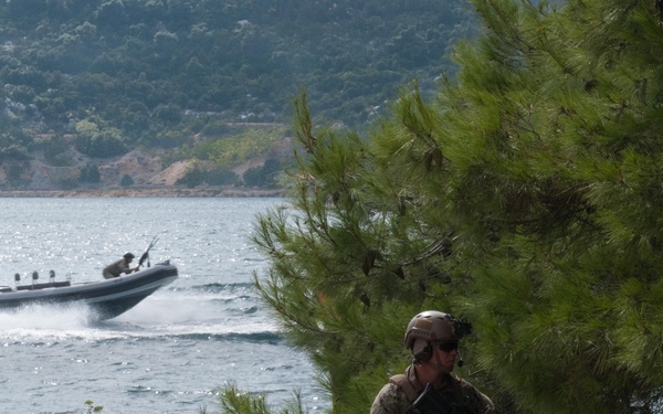 A camouflage-uniformed armed soldier is positioned in a concealed crouch behind a pine tree on a rocky shoreline, holding a rifle in a tactical stance. In the background, a rigid inflatable boat (RIB) with at least two personnel aboard is moving at high speed across the water. The scene appears to depict a military exercise or operation in a coastal/maritime environment.