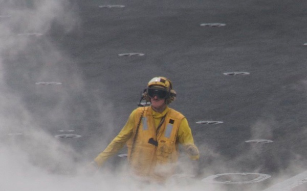 Two personnel are standing on what appears to be an aircraft carrier flight deck surrounded by dense steam or smoke. The individual in the foreground wears a tan/brown vest and yellow helmet marked with the number '3', while the person in the background wears a full yellow jersey and yellow helmet with hearing protection. The steam likely originates from a catapult launch system, which is standard on carrier flight decks.