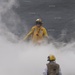 Two personnel are standing on what appears to be an aircraft carrier flight deck surrounded by dense steam or smoke. The individual in the foreground wears a tan/brown vest and yellow helmet marked with the number '3', while the person in the background wears a full yellow jersey and yellow helmet with hearing protection. The steam likely originates from a catapult launch system, which is standard on carrier flight decks.