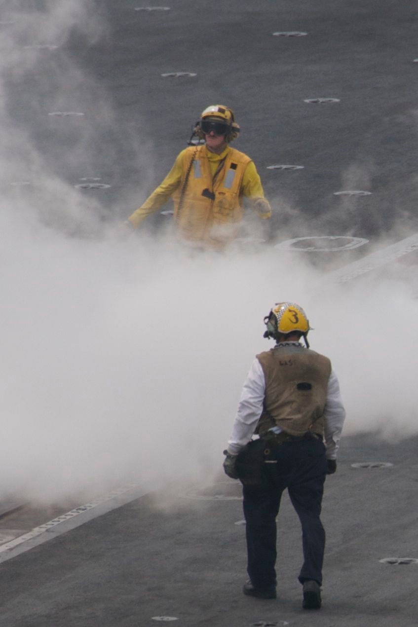 Two personnel are standing on what appears to be an aircraft carrier flight deck surrounded by dense steam or smoke. The individual in the foreground wears a tan/brown vest and yellow helmet marked with the number '3', while the person in the background wears a full yellow jersey and yellow helmet with hearing protection. The steam likely originates from a catapult launch system, which is standard on carrier flight decks.