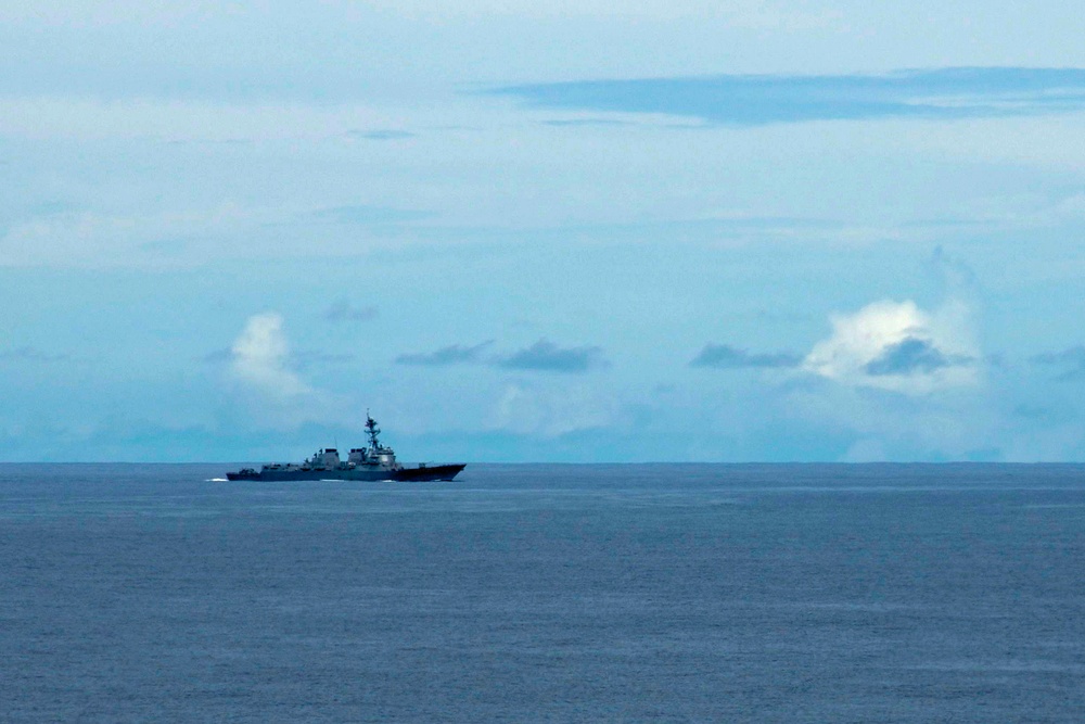 A naval warship, consistent with an Arleigh Burke-class guided-missile destroyer, is underway on open ocean under a partly cloudy blue sky. The vessel appears to be traveling at moderate speed, visible by the white wake generated at its bow and stern. The ship is photographed from a distance, placing it small against the expansive seascape.
