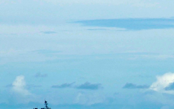 A naval warship, consistent with an Arleigh Burke-class guided-missile destroyer, is underway on open ocean under a partly cloudy blue sky. The vessel appears to be traveling at moderate speed, visible by the white wake generated at its bow and stern. The ship is photographed from a distance, placing it small against the expansive seascape.