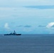 A naval warship, consistent with an Arleigh Burke-class guided-missile destroyer, is underway on open ocean under a partly cloudy blue sky. The vessel appears to be traveling at moderate speed, visible by the white wake generated at its bow and stern. The ship is photographed from a distance, placing it small against the expansive seascape.