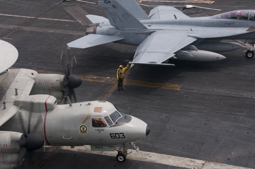 The image shows a U.S. Navy aircraft carrier flight deck with multiple aircraft and deck crew. In the foreground is a Grumman E-2 Hawkeye (bureau number 603) with spinning propellers, while an F/A-18 Hornet or Super Hornet (numbered 104) is visible in the background. A yellow-shirted aircraft handler is directing operations between the two aircraft.