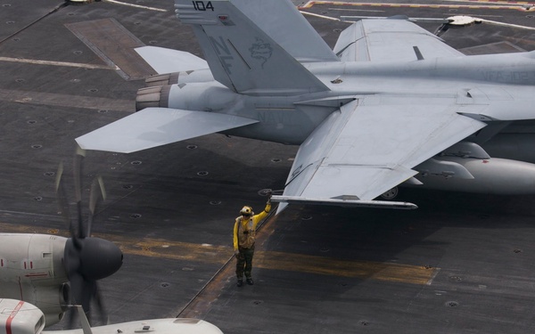 The image shows a U.S. Navy aircraft carrier flight deck with multiple aircraft and deck crew. In the foreground is a Grumman E-2 Hawkeye (bureau number 603) with spinning propellers, while an F/A-18 Hornet or Super Hornet (numbered 104) is visible in the background. A yellow-shirted aircraft handler is directing operations between the two aircraft.