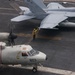 The image shows a U.S. Navy aircraft carrier flight deck with multiple aircraft and deck crew. In the foreground is a Grumman E-2 Hawkeye (bureau number 603) with spinning propellers, while an F/A-18 Hornet or Super Hornet (numbered 104) is visible in the background. A yellow-shirted aircraft handler is directing operations between the two aircraft.
