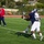 A football practice or game is taking place on a green artificial turf field. Two players in navy blue uniforms are competing for a football in mid-air, with one player attempting to catch or deflect the pass. A coach or official in a red sweatshirt and navy pants stands on the sideline observing the play.