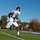 A football player wearing jersey number 8 in white uniform is running at full speed along the sideline of a turf football field during what appears to be a practice session. The low-angle shot captures the athlete mid-stride, emphasizing speed and athleticism. The setting appears to be a high school or college-level facility with a goal post visible in the background.