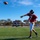 A football player wearing jersey number 7 in a red uniform has just released a pass on a well-maintained artificial turf field at Fullerton College. The image captures the ball mid-air after the throw, with other players and coaches visible in the background. The scene appears to be a practice or warm-up session under clear blue skies.