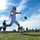 A football player in white uniform and helmet is captured mid-punt on an artificial turf field, with the ball visible just in front of his kicking leg. The low-angle shot dramatically frames the player against a bright blue sky with wispy clouds. Multiple players, coaches, and spectators are visible in the background along the sideline.