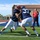 Two football players in navy blue uniforms and white pants are engaged in a blocking/tackling drill on an artificial turf field. One player wears a red face mask helmet, while the other wears jersey number 37. A coach in a red shirt observes in the background near blue training dummies.